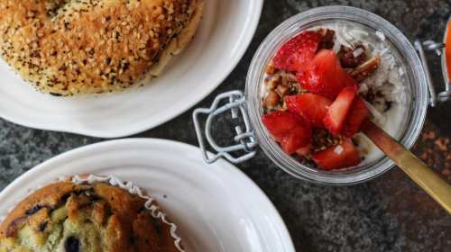 A plate with a sesame bagel and a muffin, alongside a jar of yogurt topped with strawberries and granola.