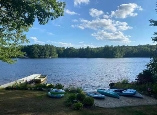 A serene lake view with kayaks and inflatable floats on the shore, surrounded by trees and a clear blue sky.