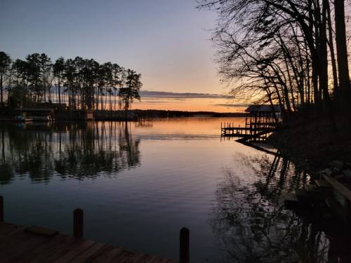 Sunset over a calm lake, with trees lining the shore and reflections on the water. A dock is visible in the foreground.