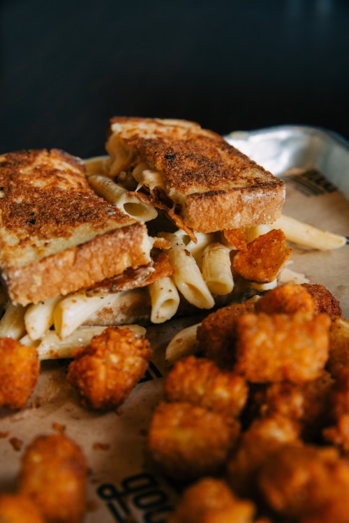 A close-up of a grilled cheese sandwich with pasta and crispy tater tots on a tray.