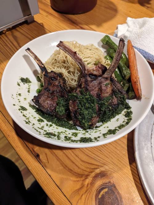 A plate of grilled lamb chops with green sauce, served with noodles and assorted vegetables on a wooden table.