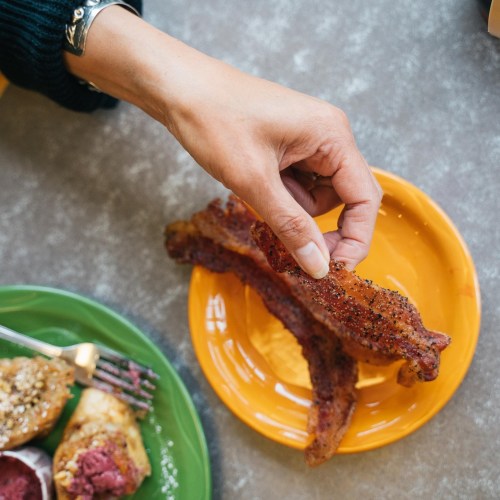 A hand reaches for crispy bacon on an orange plate, with a green plate of food in the foreground.