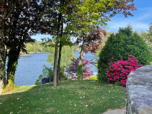 A serene lakeside view with vibrant flowers, lush trees, and calm water under a clear blue sky.