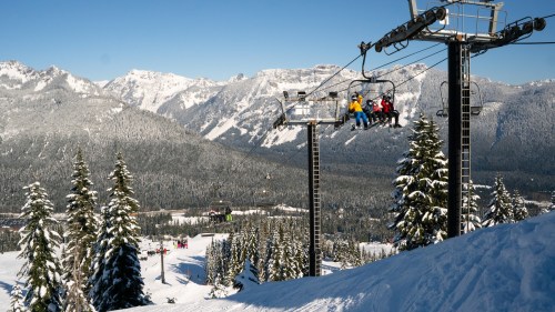 Skiers on a chairlift above a snowy mountain landscape with tall trees and clear blue skies.