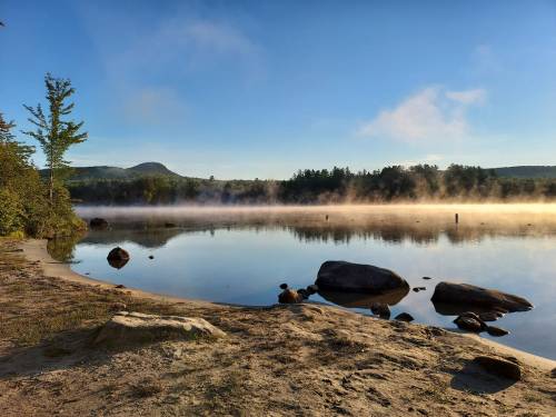A serene lake at dawn, with mist rising from the water and rocky shores surrounded by trees and distant hills.