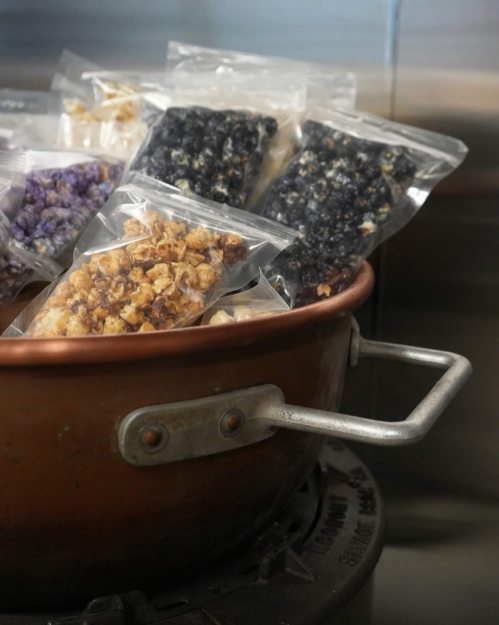 A copper pot filled with bags of colorful popcorn, including purple and caramel varieties, on a stovetop.