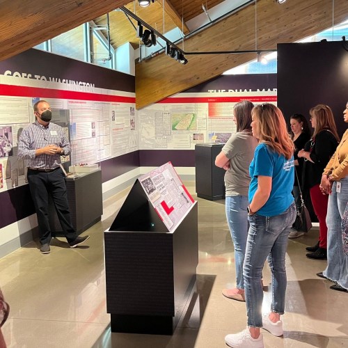 A group of people listens to a speaker in a museum exhibit with informational panels and displays.