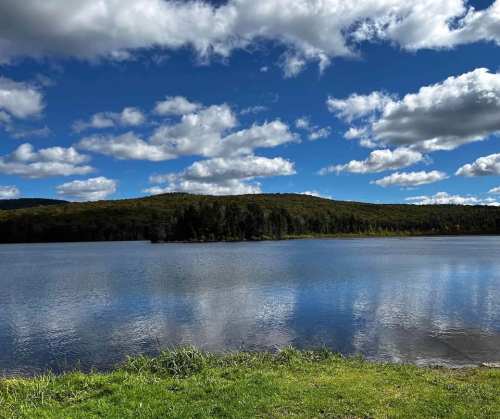 A serene lake surrounded by green hills and a blue sky with fluffy white clouds reflecting on the water's surface.