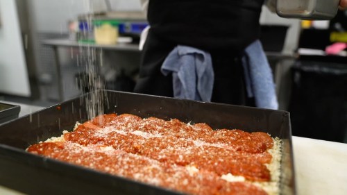 A person sprinkles seasoning over a tray of pizza with pepperoni and cheese in a kitchen setting.