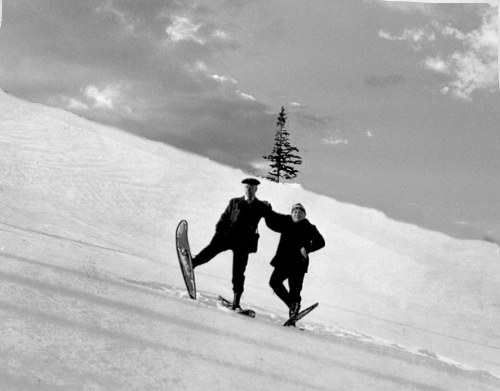 Two people on skis pose playfully on a snowy slope, with a lone tree in the background under a cloudy sky.