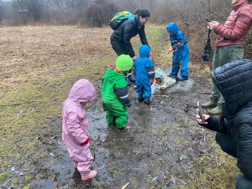 Children in colorful rain gear play in a muddy area while adults supervise and take photos in a natural setting.