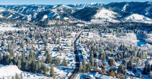 Aerial view of a snowy town surrounded by mountains and forests, with a winding road and houses blanketed in snow.