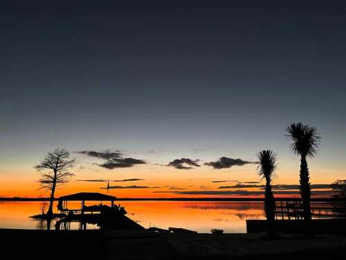 Silhouette of palm trees and a dock against a vibrant sunset over calm water, with darkening sky and clouds.