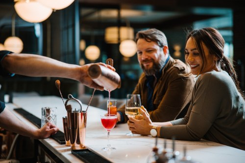 A bartender pours a cocktail for a smiling woman at a bar, while a man looks on, enjoying his drink.
