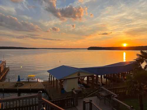 Sunset over a calm lake, with docks and a blue-roofed building in the foreground, surrounded by trees and clouds.