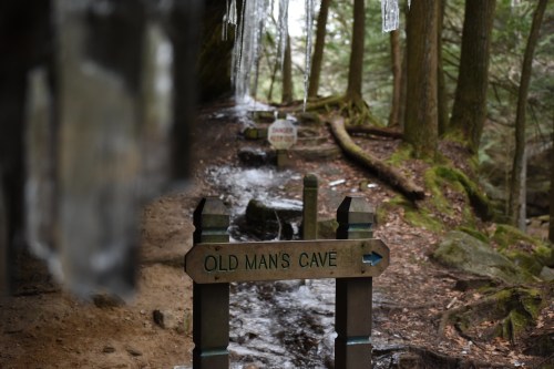 A wooden sign points to "Old Man's Cave" along a forested trail with icicles and a stream in the background.