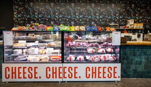 A cheese display case filled with various cheeses, with a colorful backdrop and a sign that reads "CHEESE. CHEESE. CHEESE."