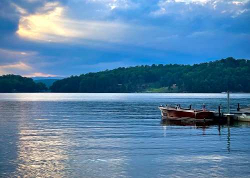 A calm lake at sunset with a small boat docked and lush green hills in the background under a cloudy sky.