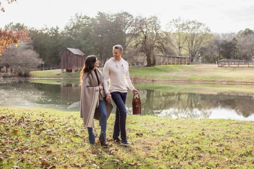 A couple walks hand in hand by a serene pond, surrounded by trees and a rustic building in the background.