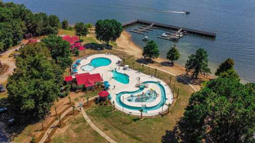 Aerial view of a lakeside resort featuring two swimming pools, green trees, and a dock with boats.