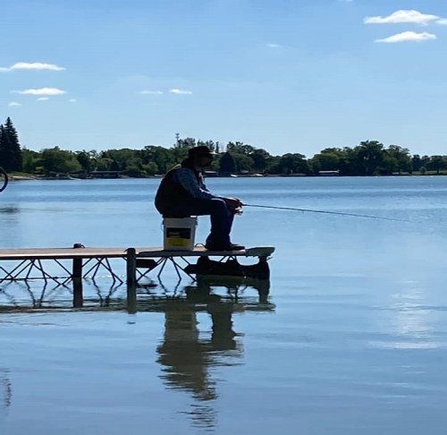 A person fishing from a dock by a calm lake on a sunny day, surrounded by trees and blue skies.