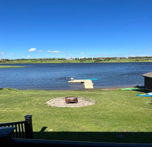 A serene lakeside view with a dock, green grass, and a fire pit under a clear blue sky.