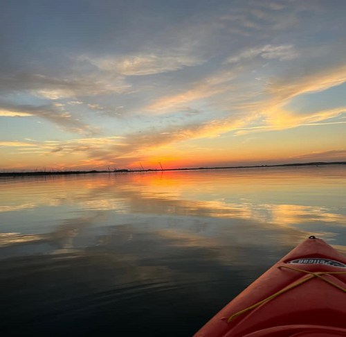 A kayak on calm water reflects a vibrant sunset with colorful clouds and a serene landscape in the background.