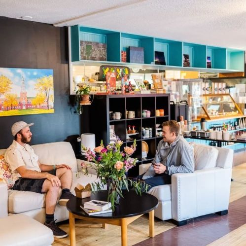 Two men sit in a cozy café, chatting with a table of flowers and snacks between them, surrounded by colorful decor.