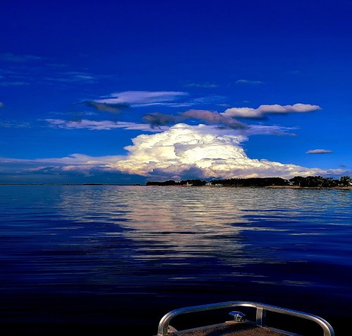 A serene lake scene with calm waters reflecting fluffy clouds and a blue sky, bordered by a distant shoreline.