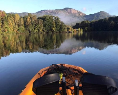 A kayak on calm water, surrounded by trees and mountains under a clear blue sky.