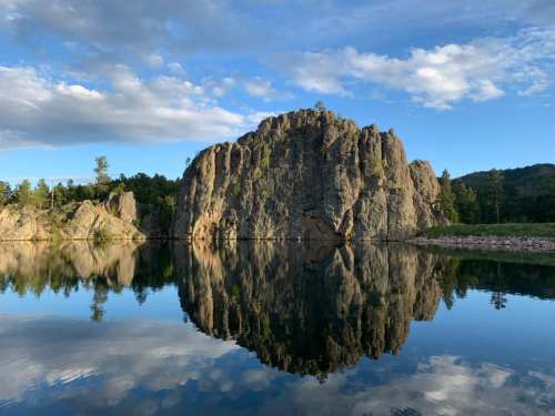 A serene landscape featuring a rocky cliff reflected in calm water, surrounded by trees and a blue sky with clouds.
