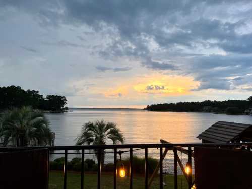 A serene lakeside view at sunset, with clouds and reflections on the water, framed by trees and a balcony railing.