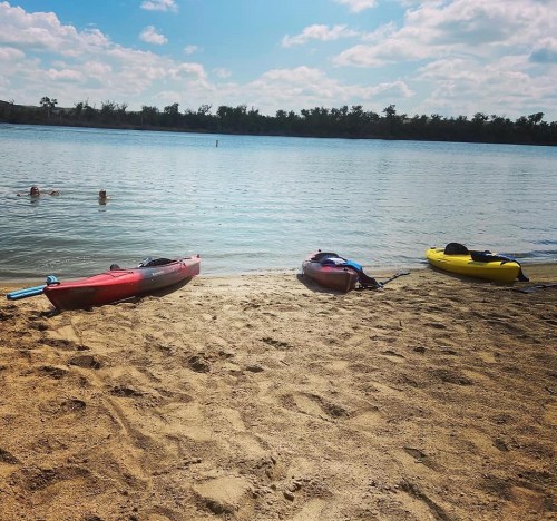 Two people swim in a calm lake, with red and yellow kayaks resting on the sandy shore under a sunny sky.