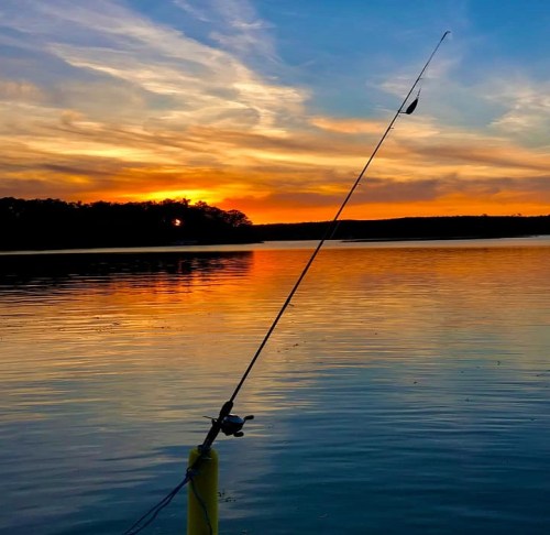 A fishing rod stands against a vibrant sunset over a calm lake, reflecting warm colors on the water's surface.