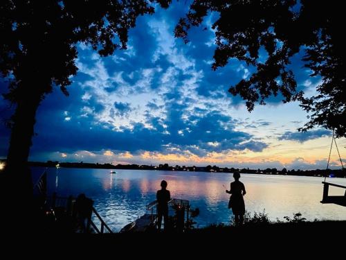 Silhouettes of two people by a lake at sunset, with colorful clouds reflecting on the water.