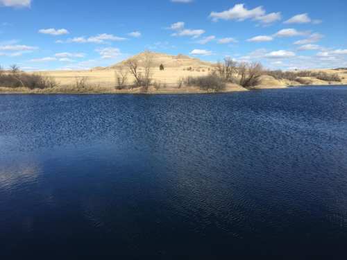 A serene lake reflects the blue sky, surrounded by rolling hills and sparse trees under a partly cloudy day.