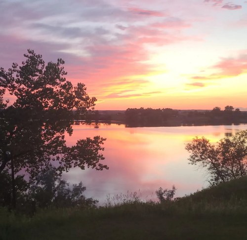 A serene lake at sunset, reflecting vibrant pink and purple skies, framed by trees on the shore.