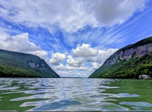 A serene lake surrounded by lush green hills under a bright blue sky with fluffy clouds.