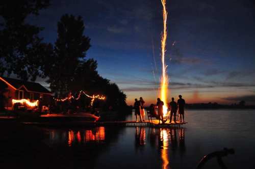 Silhouetted figures on a dock celebrate with fireworks over a calm lake at dusk, surrounded by festive lights.