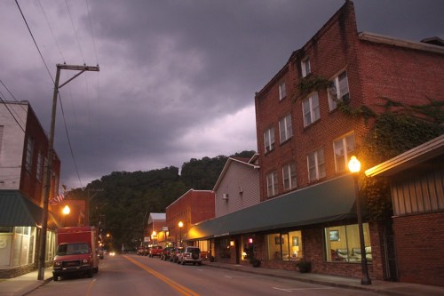 A quiet street in a small town, lined with brick buildings under a dark, cloudy sky. Streetlights glow softly.