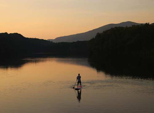A person paddleboards on a calm lake at sunset, surrounded by mountains and trees.