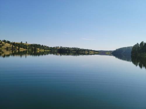A calm lake reflecting clear blue skies and surrounding green hills, creating a serene landscape.