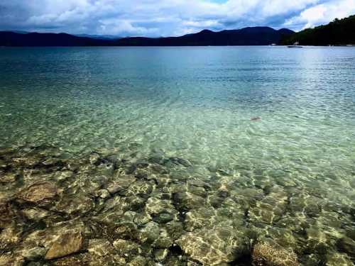 Clear water over rocky seabed, with distant mountains and cloudy skies in the background.