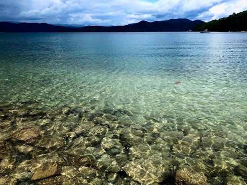 Clear water over rocky seabed, with distant mountains and cloudy skies in the background.