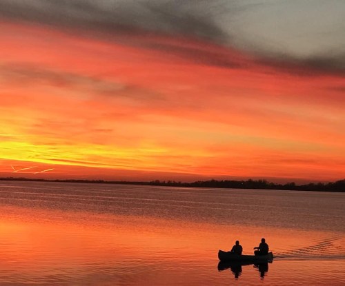 Two people in a canoe on a calm lake at sunset, surrounded by vibrant orange and pink skies.