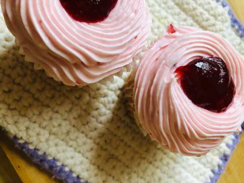 Two pink frosted cupcakes with a dollop of red jam on top, placed on a textured cream-colored surface.