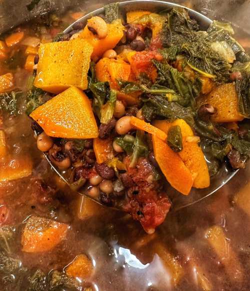 A close-up of a hearty vegetable stew with squash, greens, black-eyed peas, and tomatoes in a pot.