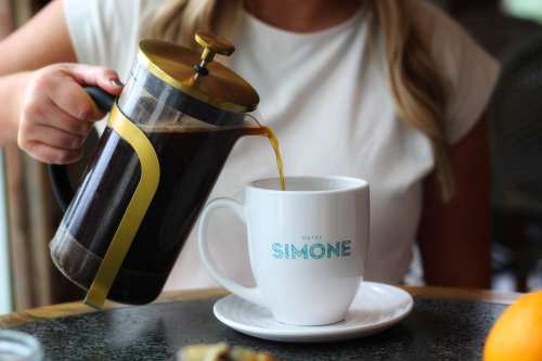 A person pours coffee from a French press into a white mug with "SIMONE" printed on it, set on a table.