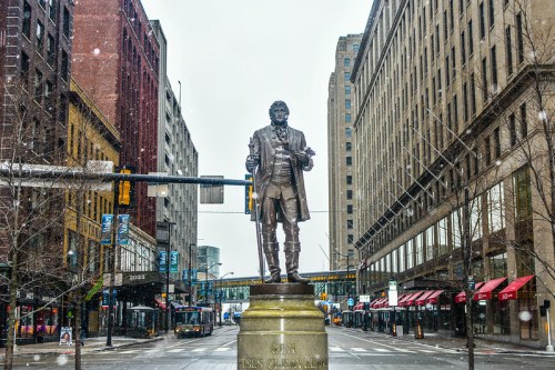 Statue of a historical figure in a snowy urban street, surrounded by buildings and empty sidewalks.