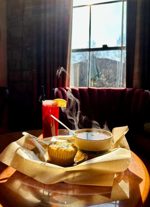 A steaming bowl of soup and a muffin on a table, with a red drink and sunlight streaming through a window.